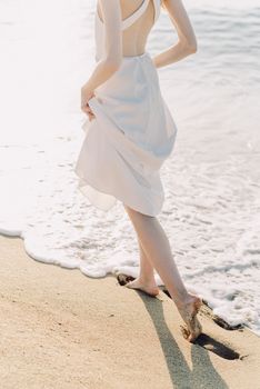 A woman in a white dress walks gracefully along a sunny beach, toes in the sand.