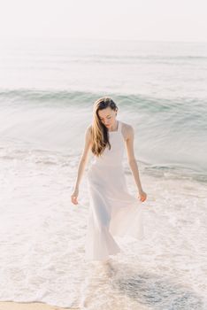 Elegant woman in a flowing white dress strolling along a sunny beach, enjoying a summer day.