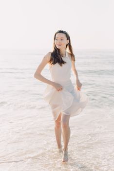 Elegant woman in a white dress joyfully walks along the serene beach at sunset.