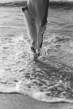 A grayscale image of an elegant woman walking through waves at the beach.