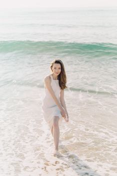 A woman in a white dress joyfully walks by the ocean shore on a sunny day.