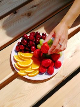 A vibrant assortment of fresh fruits on a plate with a hand reaching over wooden surface.