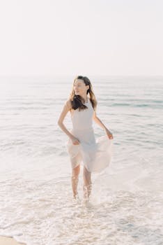 Woman in white dress walking along a serene beach during sunrise.