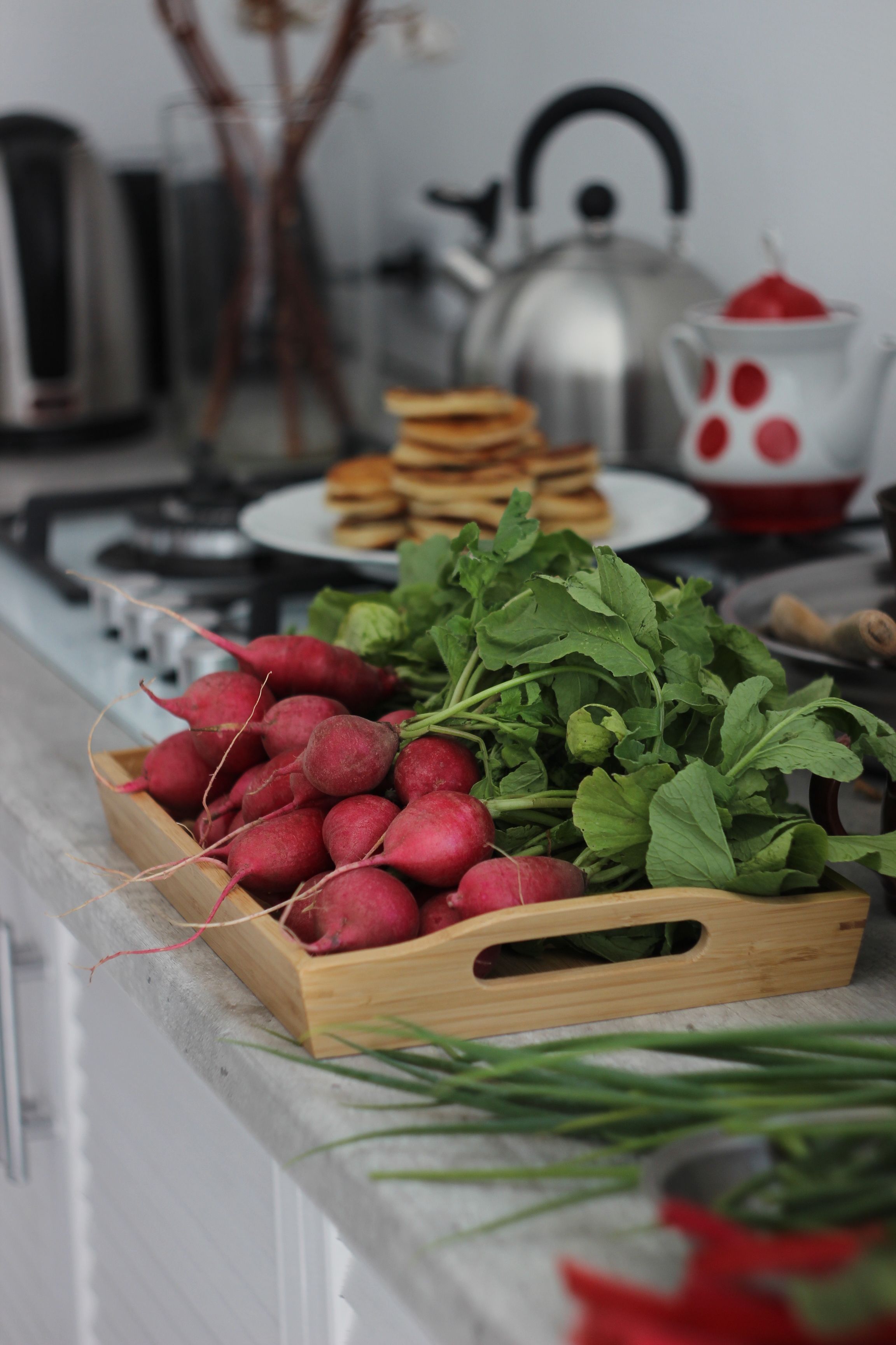 A wooden tray of fresh radishes on a kitchen counter with a stack of pancakes in the background.