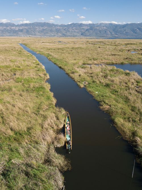 Free A serene aerial shot of a canoe on a canal in Inle Lake, surrounded by lush grasslands and mountains. Stock Photo