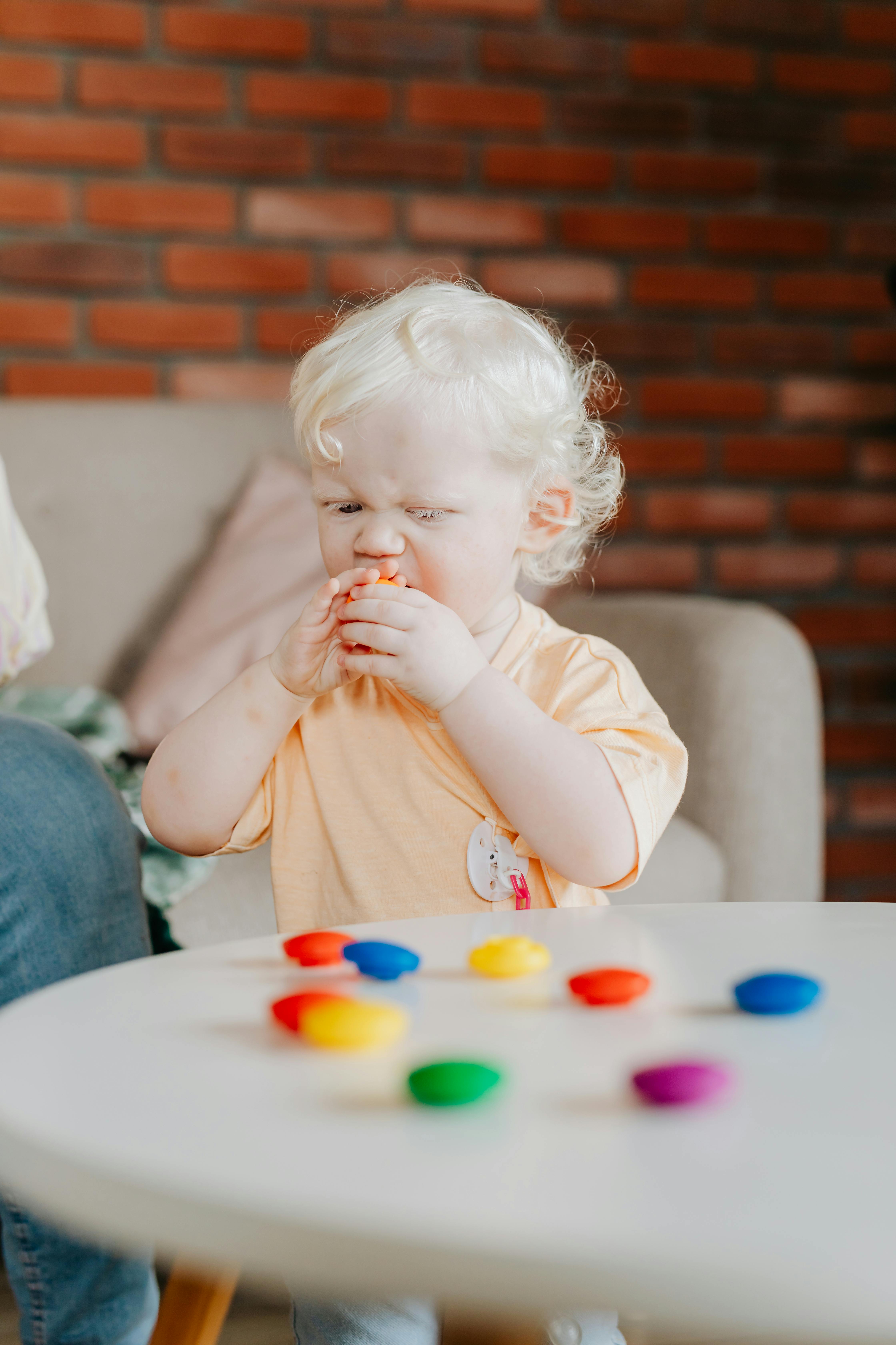 Cute child with white hair eating while standing near a table with colorful toys indoors.