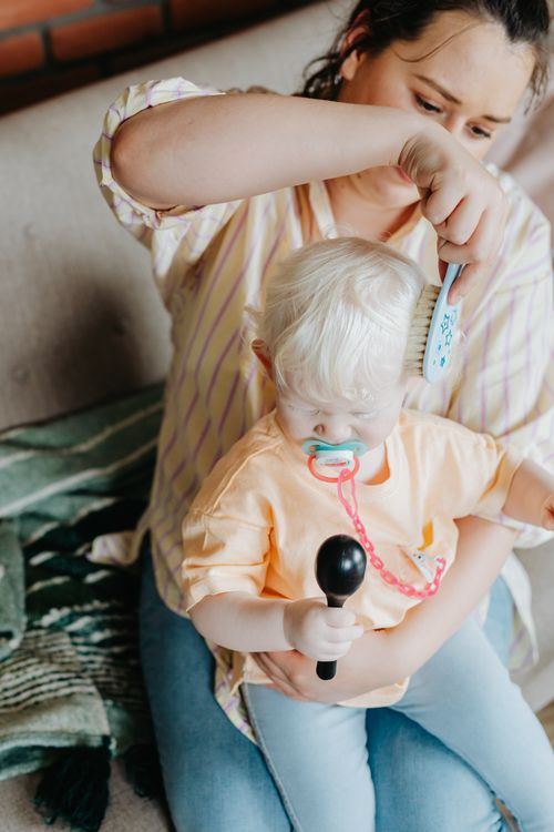 Free A mother brushes her baby's hair while they sit on a couch. The baby holds a pacifier. Stock Photo