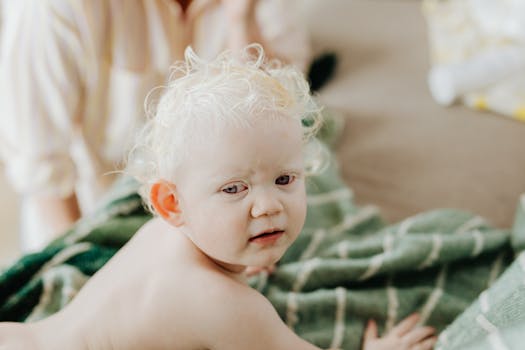 Cute baby with curly hair looking over shoulder, lying on a soft blanket indoors.