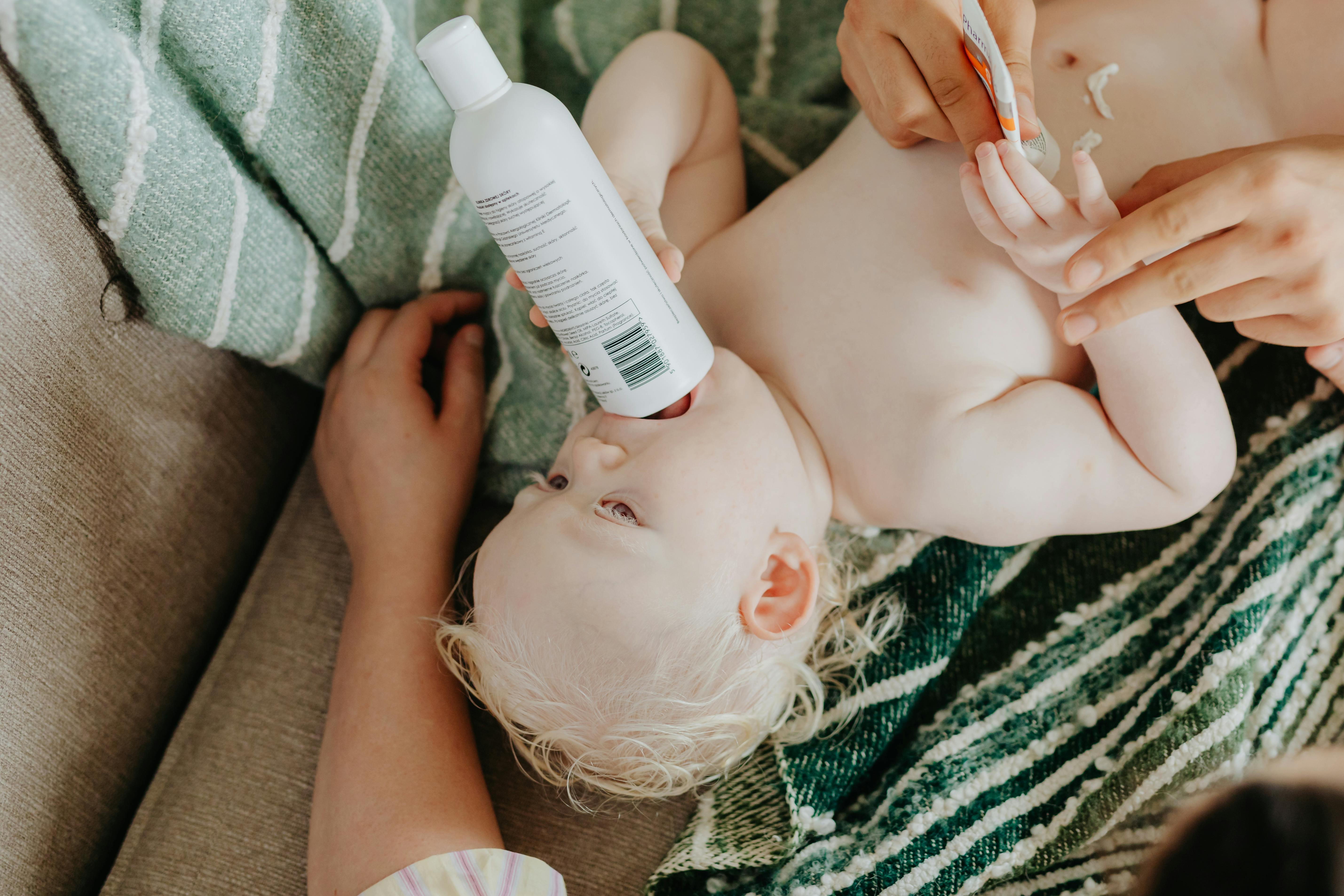 A cute baby lies comfortably while holding a lotion bottle during a skincare routine.