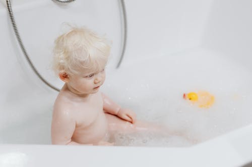 Photograph of a Child with Blond Hair Sitting in a Bathtub with Bubbles