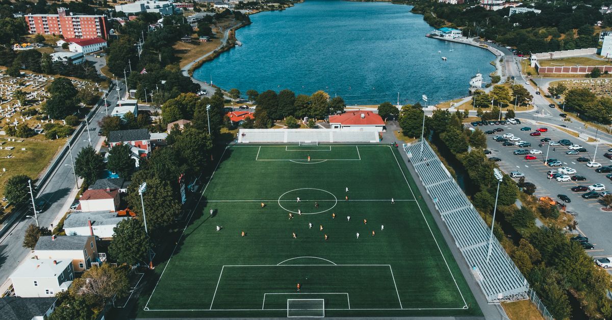 Aerial view of a soccer field by a scenic waterfront with players and surrounding cityscape.