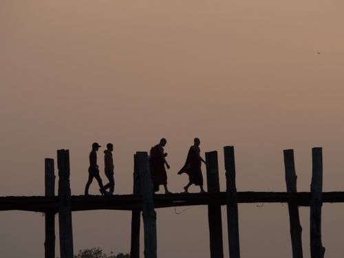 Free People crossing U Bein Bridge in Mandalay, Myanmar, during dusk, creating serene silhouettes against the sunset. Stock Photo