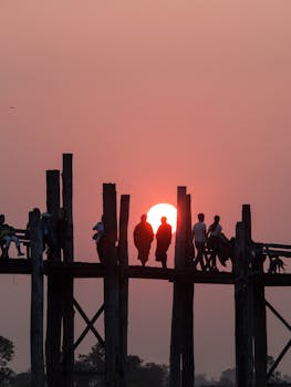 People walking on the iconic U-Bein Bridge in Mandalay during a stunning sunset, with the sun setting behind their silhouettes.