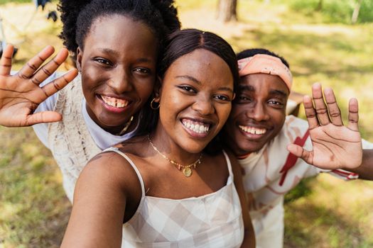 Three young friends capture a joyful outdoor selfie with bright smiles and waving hands.