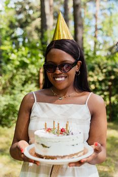 A joyful woman wearing a party hat holds a birthday cake in a sunlit garden.