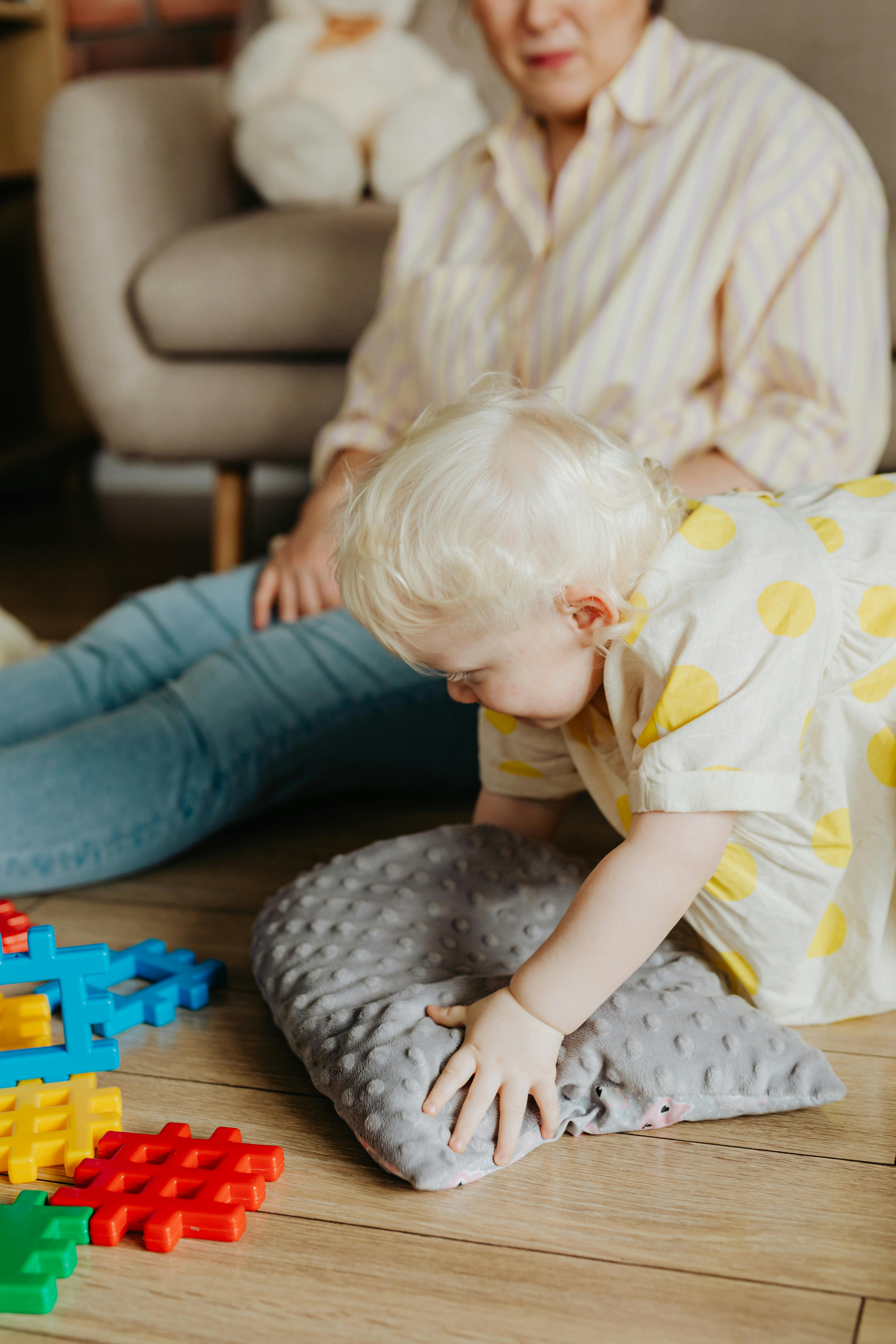 A cute toddler with light hair engaging with colorful toys indoors, supervised by an adult.