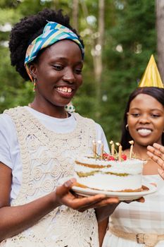 Two women celebrating a birthday outdoors, one holding a cake with lit candles, both smiling.