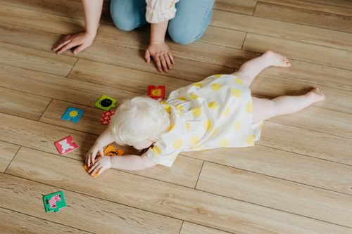 Free A baby in a polka dot dress plays with colorful cards on a wooden floor indoors. Stock Photo
