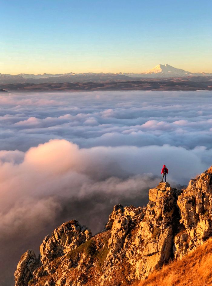 A lone hiker stands atop a cliff at sunrise, overlooking clouds — photo by Artem Meletov on Pexels