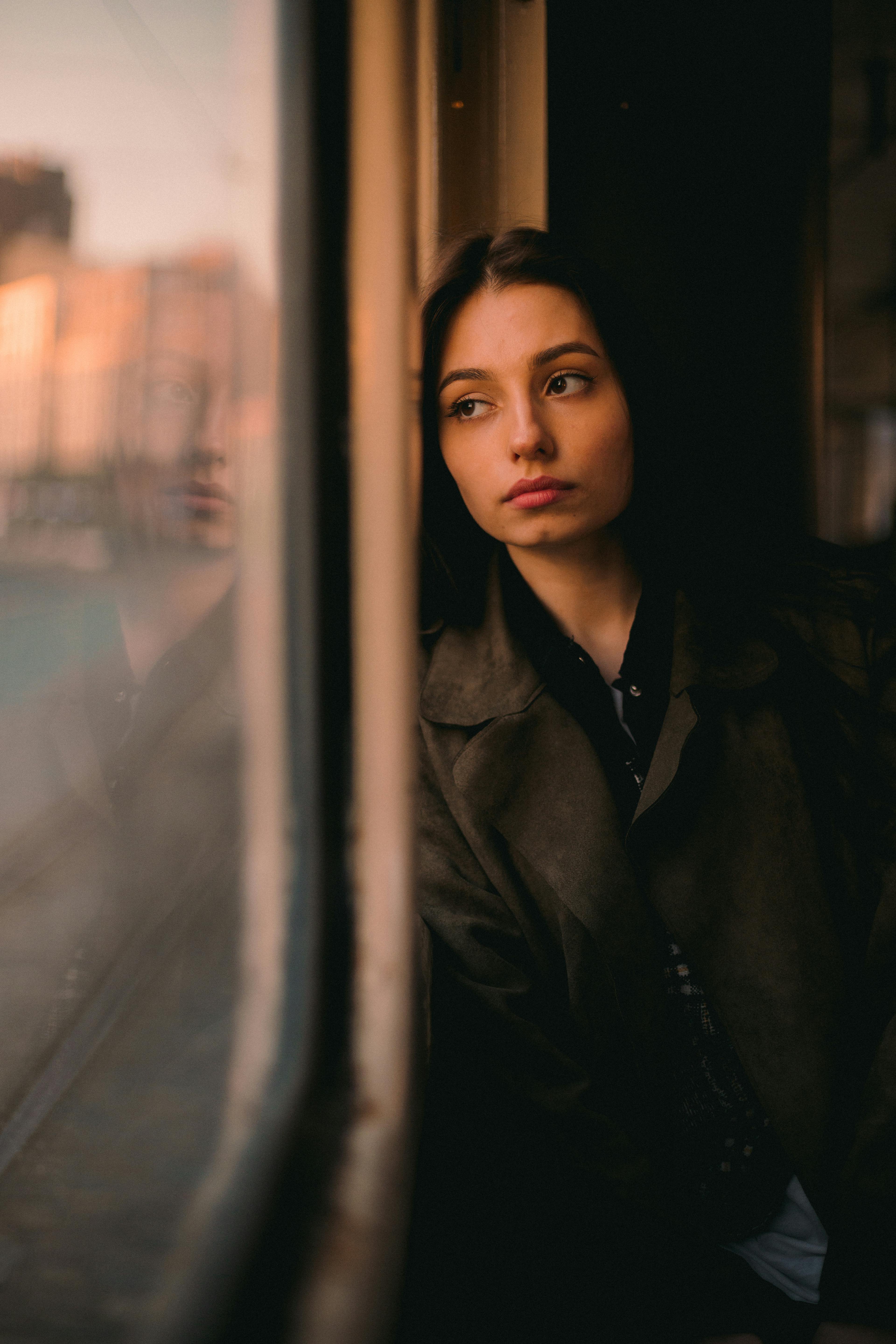 Young woman leaning against a window, gazing outside with a contemplative look.