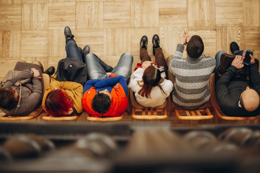 Top view of adults sitting in a row on wooden chairs on a parquet floor indoors.