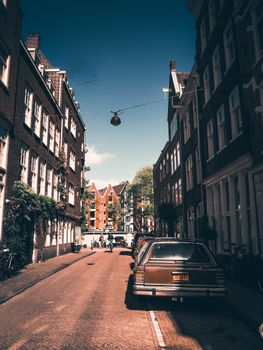 Atmospheric street scene in Amsterdam with parked cars and traditional buildings under a blue sky.