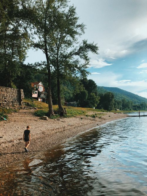 Free A child walks along a scenic lakeside shore with trees and mountains in Zolnoye, Russia. Stock Photo