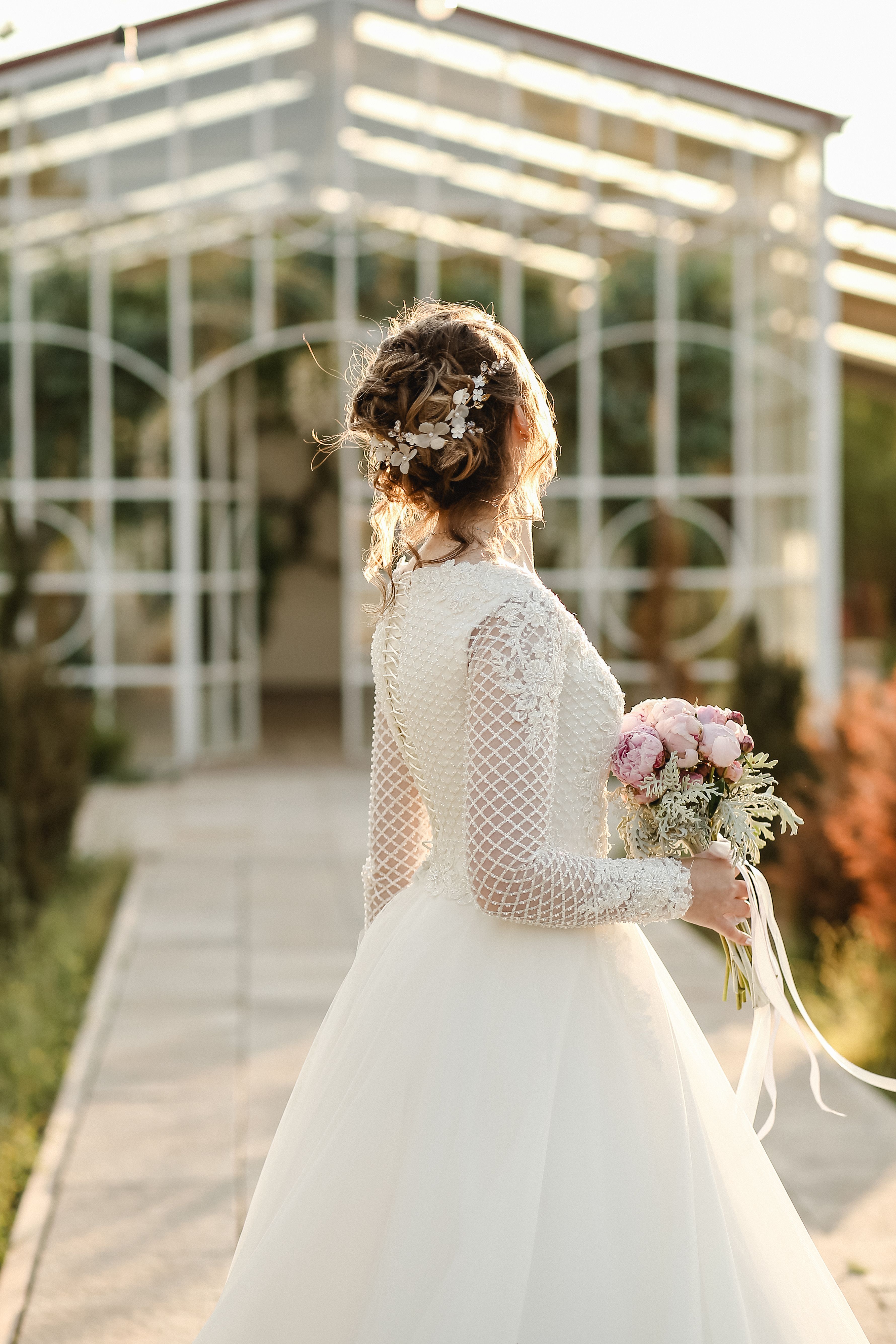 A bride in a lace gown holding a pink bouquet with a greenhouse backdrop at an outdoor wedding.