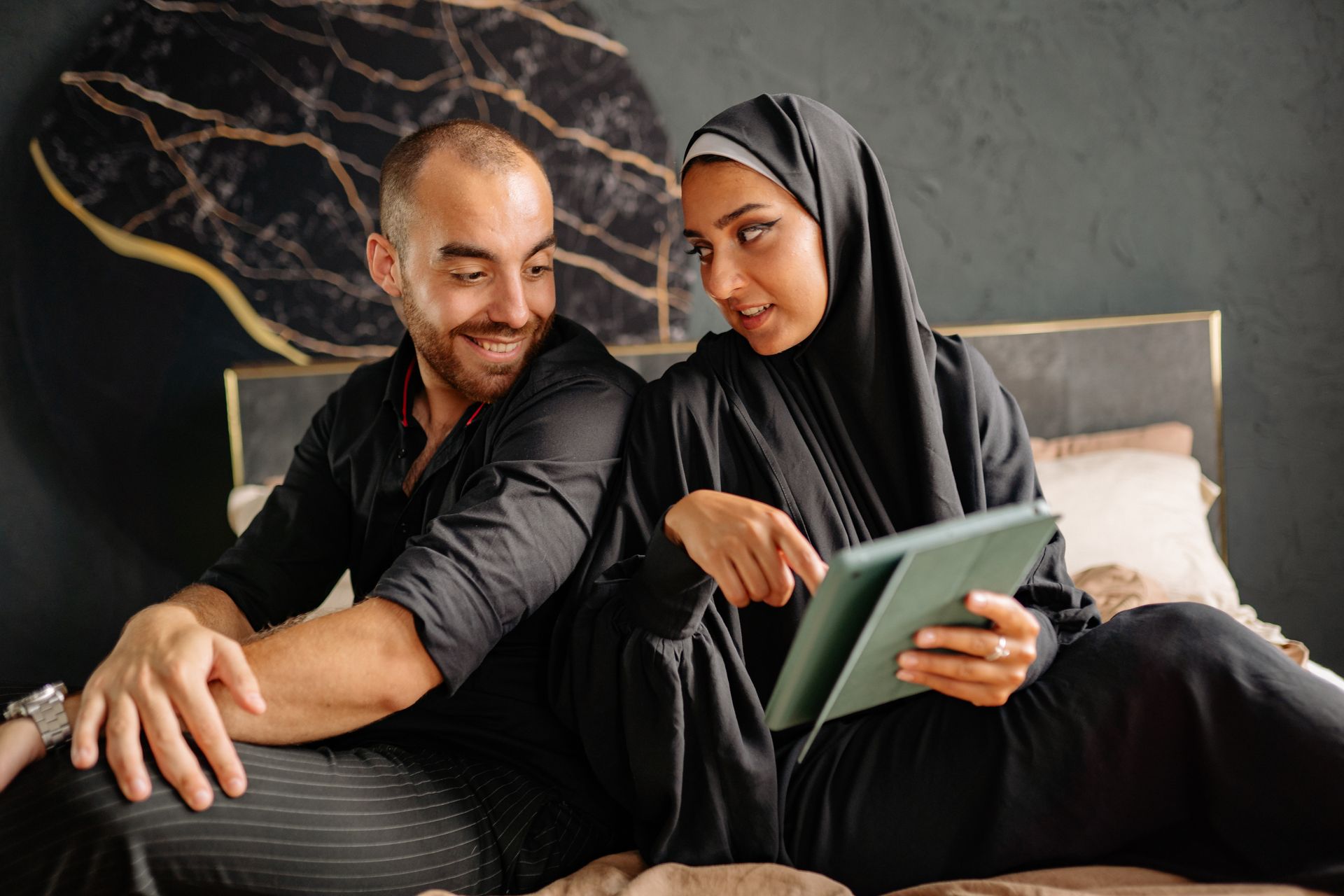 A diverse couple sitting on a bed and discussing something on a tablet indoors.