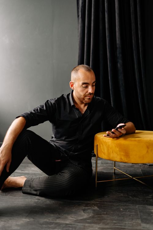 Free Man in casual black attire sits on the floor indoors, using a smartphone next to a yellow stool. Stock Photo