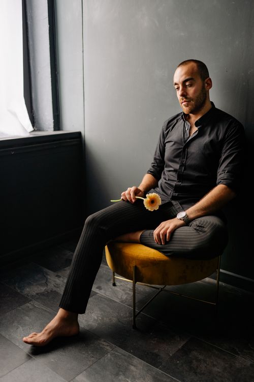 Free A thoughtful man sits barefoot, holding a flower in a dimly lit room. Stock Photo
