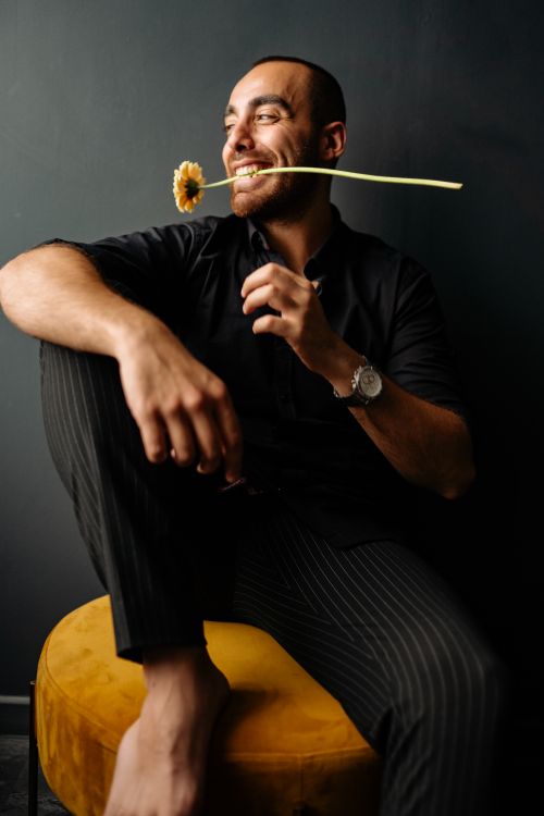 Free A joyful man sits barefoot on a chair, holding a flower in his mouth, exuding happiness and style. Stock Photo