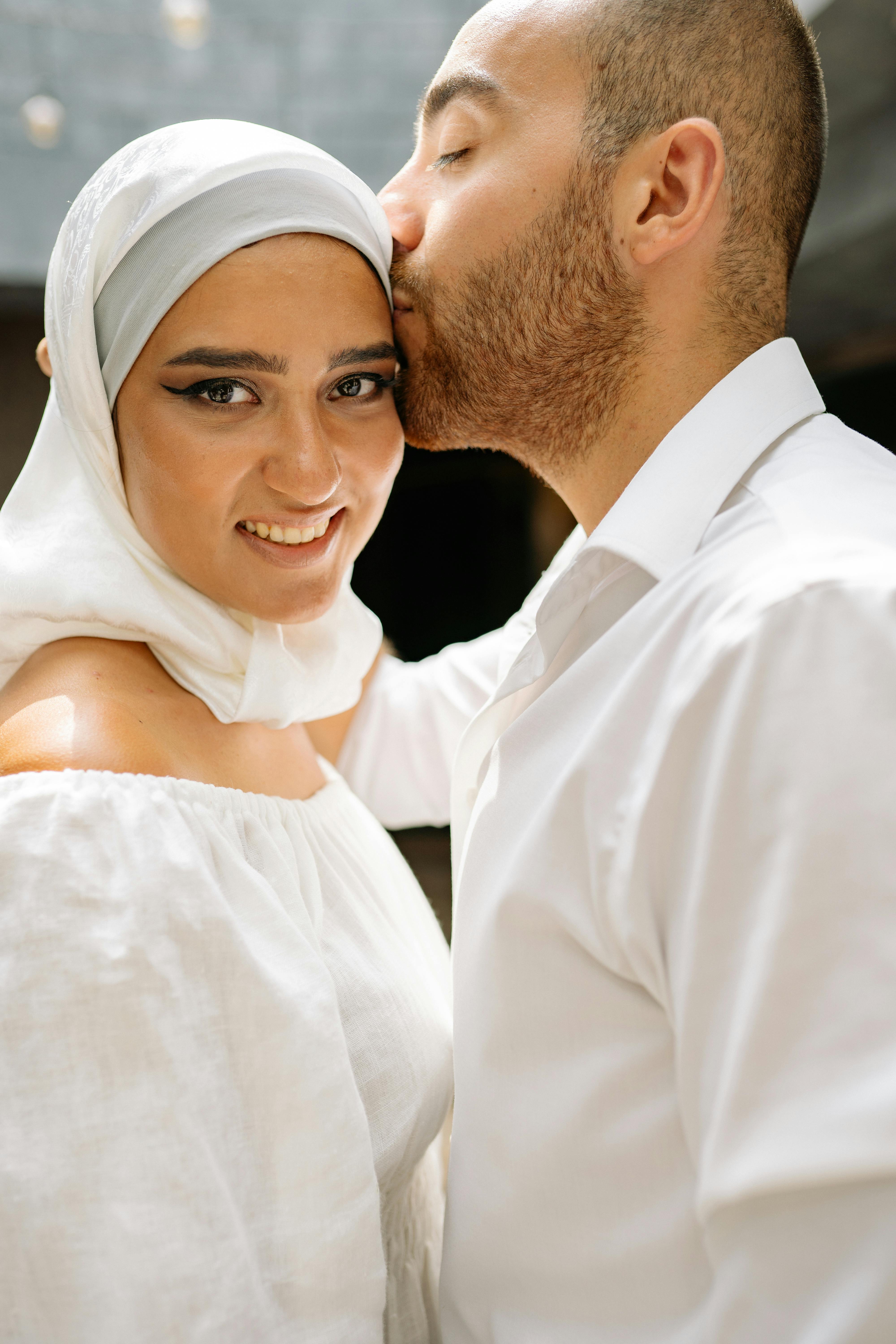 A loving couple shares a tender moment with a kiss and smile, showcasing elegance in white attire.