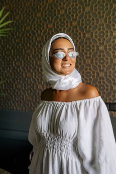 Stylish woman wearing a white hijab and dress, smiling indoors with sunglasses.
