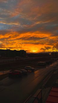 A vibrant city sunset with dramatic clouds and silhouetted cars on a wet road.