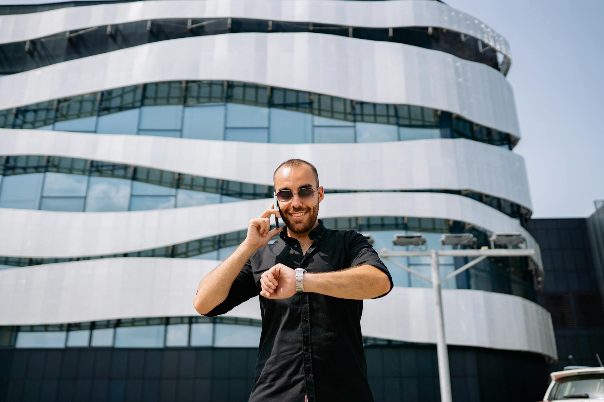 Confident man on phone call checking wristwatch in front of modern building.