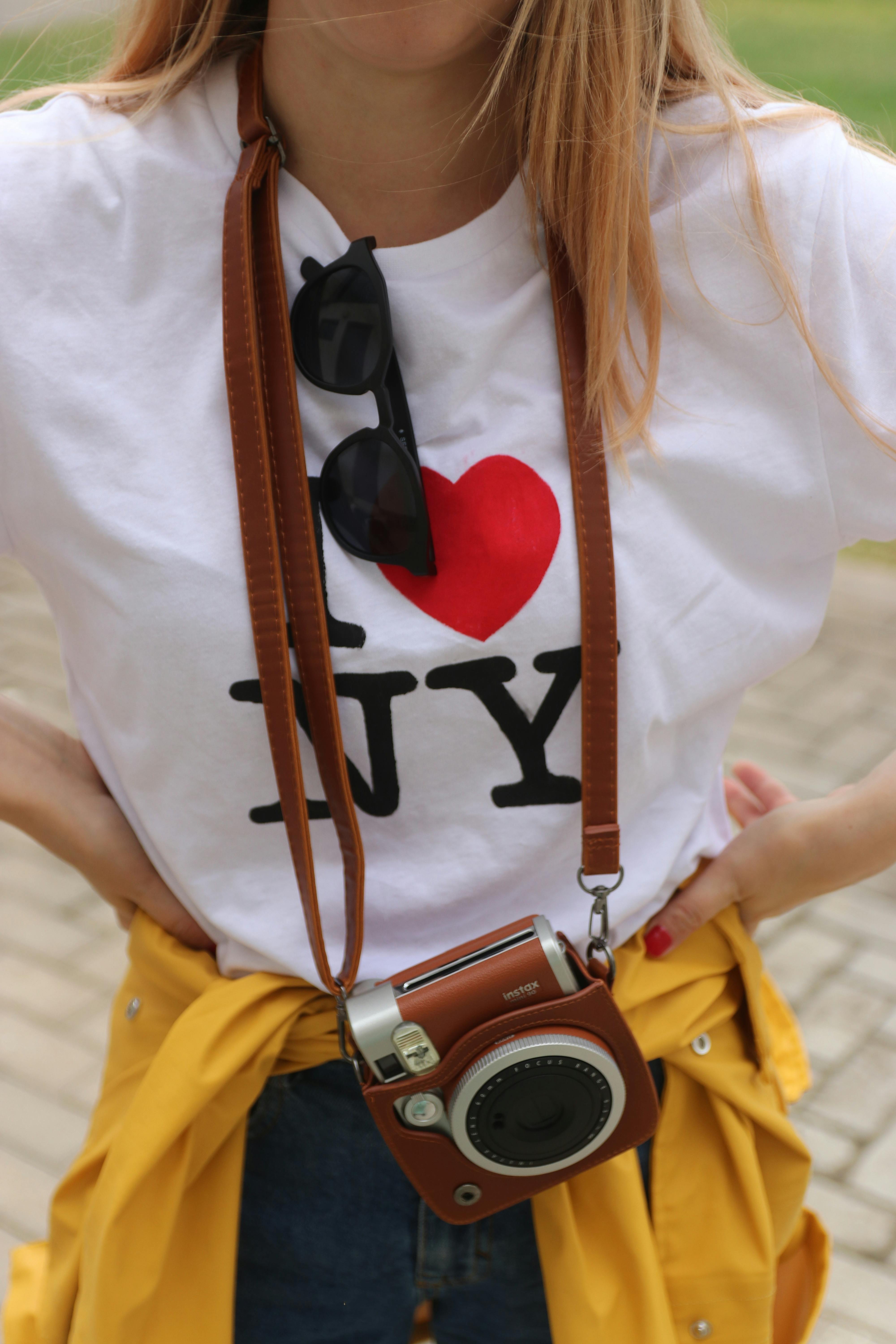 Modern woman in 'I ♥ NY' t-shirt with vintage camera outdoors, balancing casual and fashionable style.