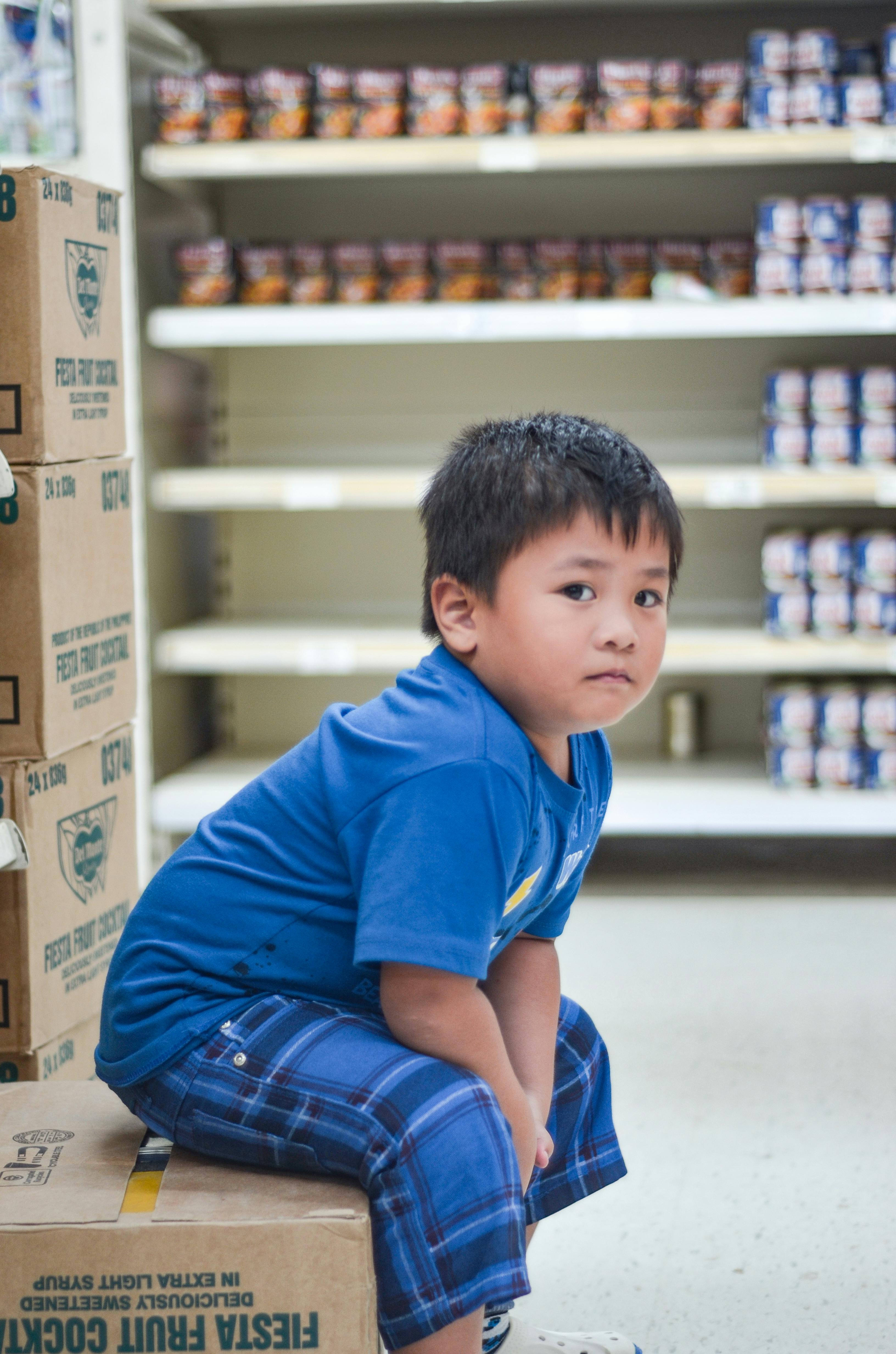 一个小男孩穿着蓝色衣服，坐在杂货店过道的盒子上，四周都是货架