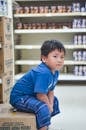 一个小男孩穿着蓝色衣服，坐在杂货店过道的盒子上，四周都是货架