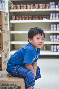 一个小男孩穿着蓝色衣服，坐在杂货店过道的盒子上，四周都是货架