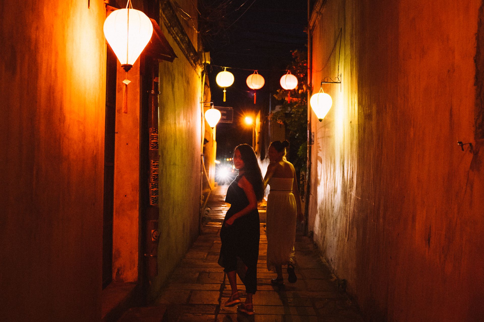 Two women walking under glowing lanterns in a narrow, dimly lit street.