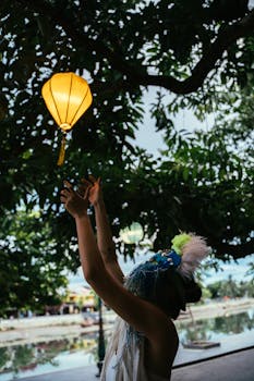 A woman releases a glowing lantern during a Taiwanese festival under lush trees.