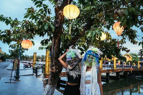 People in Ethnic Masks Standing by a Tree with Paper Lanterns