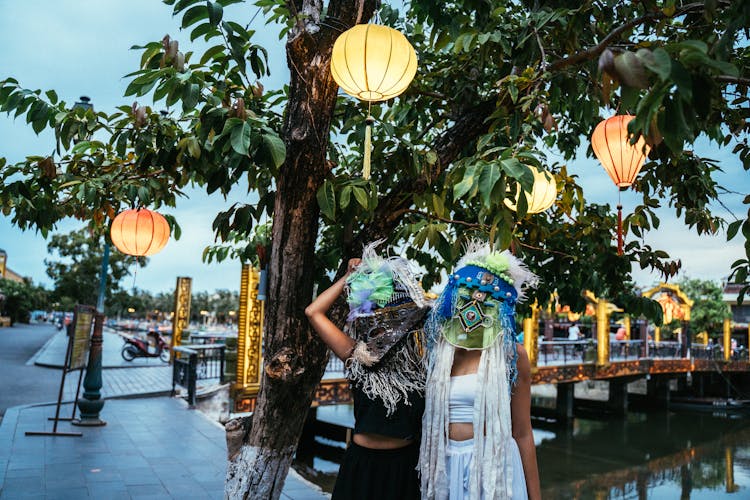 People In Ethnic Masks Standing By A Tree With Paper Lanterns