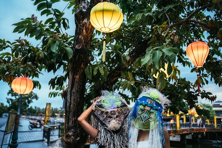 People In Ethnic Masks Standing By A Tree With Paper Lanterns