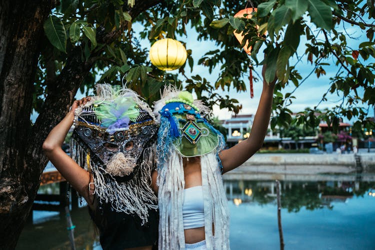 People In Ethnic Masks Standing By A Tree With Paper Lanterns
