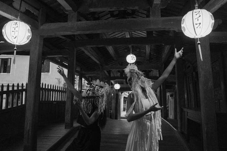 Grayscale Photo Of People In Ethnic Masks In A Footbridge With Paper Lanterns