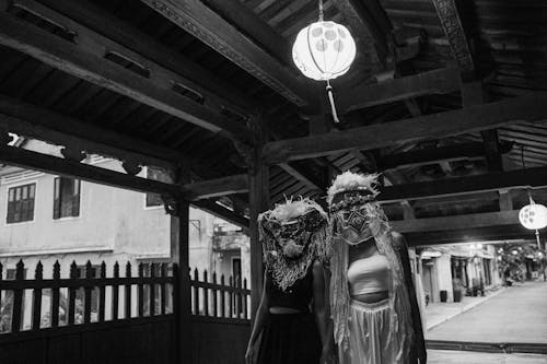 Grayscale Photo of People in Ethnic Masks Standing under a Paper Lantern