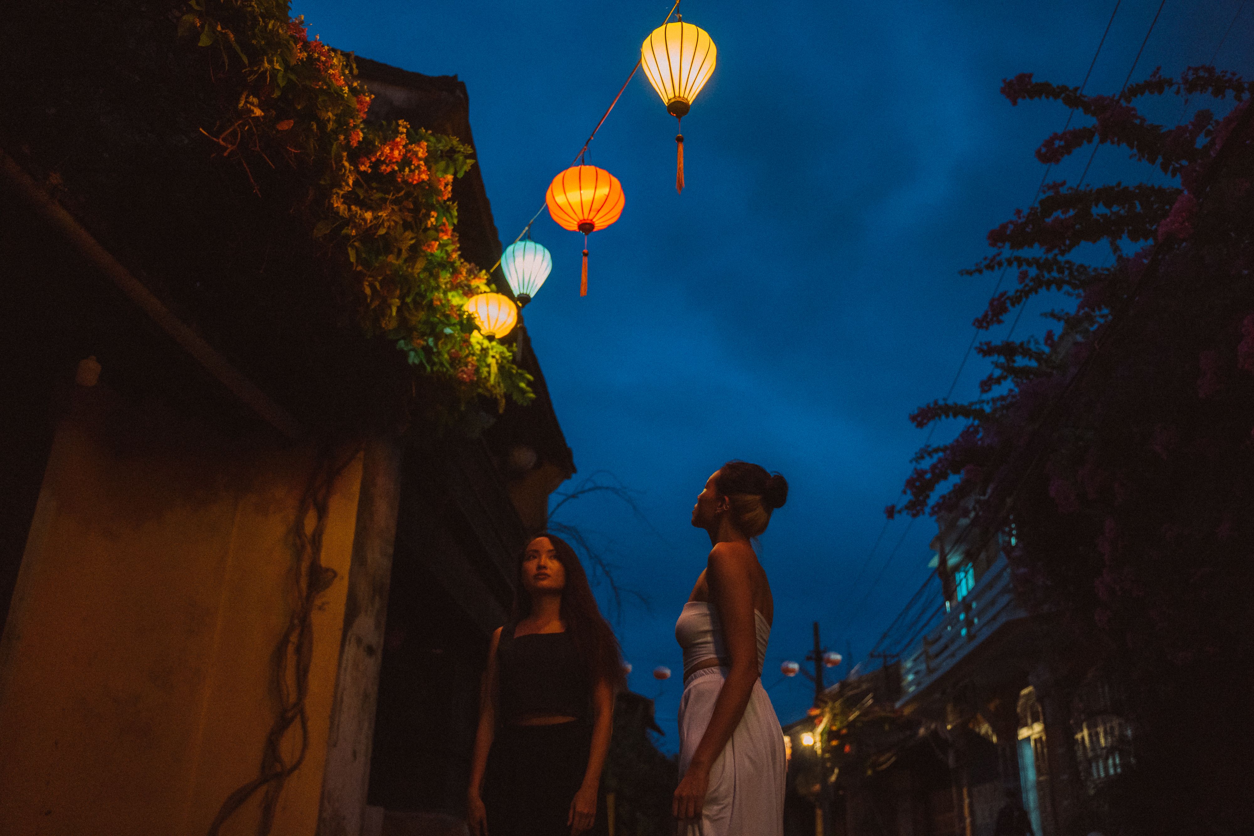 Two Asian women standing under colorful lanterns in a traditional urban alley at night.