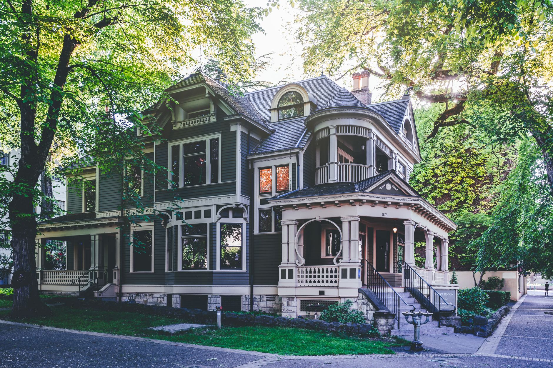 A large, historic, two-story house with dark siding and ornate white trim nestled among trees.